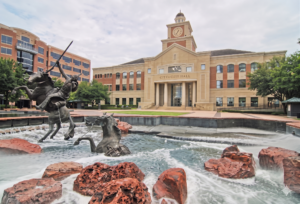 A large bronze sculpture of a Native American warrior on horseback in a rushing water fountain in front of a grand city hall building.